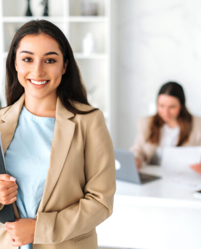 Positive successful pretty indian or arabian business lady, secretary, company executive, financial manager, in a stylish suit stand in a modern office, holding documents, looking and smiles at camera
