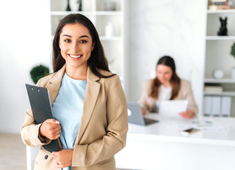 Positive successful pretty indian or arabian business lady, secretary, company executive, financial manager, in a stylish suit stand in a modern office, holding documents, looking and smiles at camera